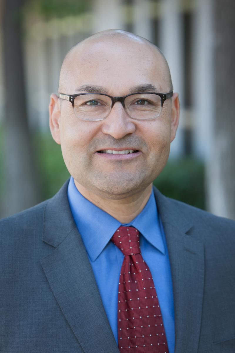 Head and shoulders portrait of a smiling, clean-shaven, bespectacled Latinx man in a gray business suit.