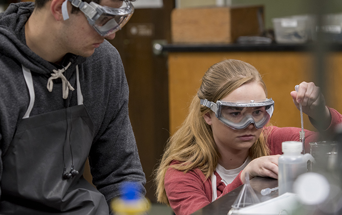 Two students wearing safety goggles as they handle chemistry lab materials