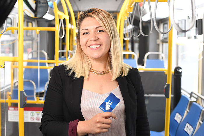 Waist-up image of a smiling blonde woman in black jacket, standing inside a municipal bus, holding a Clipper Card up to the camera.