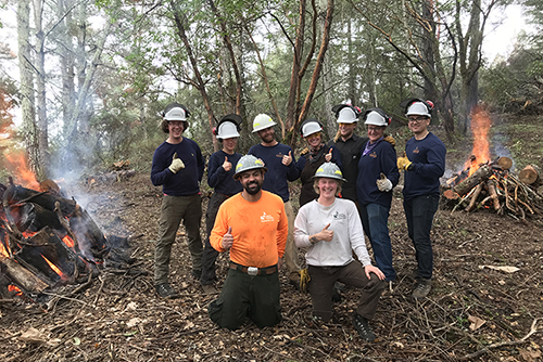 Nine people in workgear posing amid small controlled burn piles, smiling and giving a thumbs up