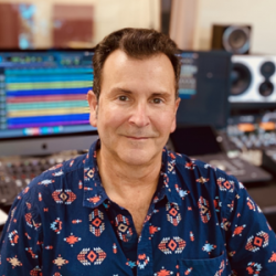 Head and shoulders portrait shot of Len Hayes, a smiling man in a colorful button down short, seated in front of a computer screen.