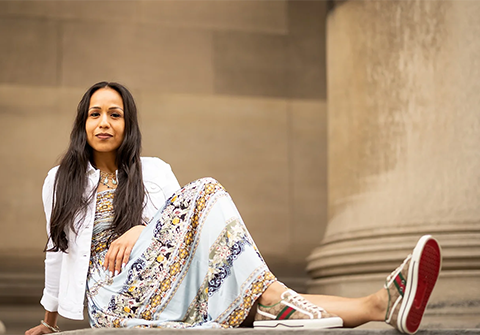 Casual shot of Dr. Gina Garcia in a floral dress, white sweater and sneakers, seated on the ground in front of a large sandstone pillar.