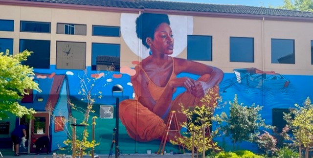 Building exterior; full-height mural of a brown-skinned woman dressed in orange, surrounded by blue water and haloed by a full moon.