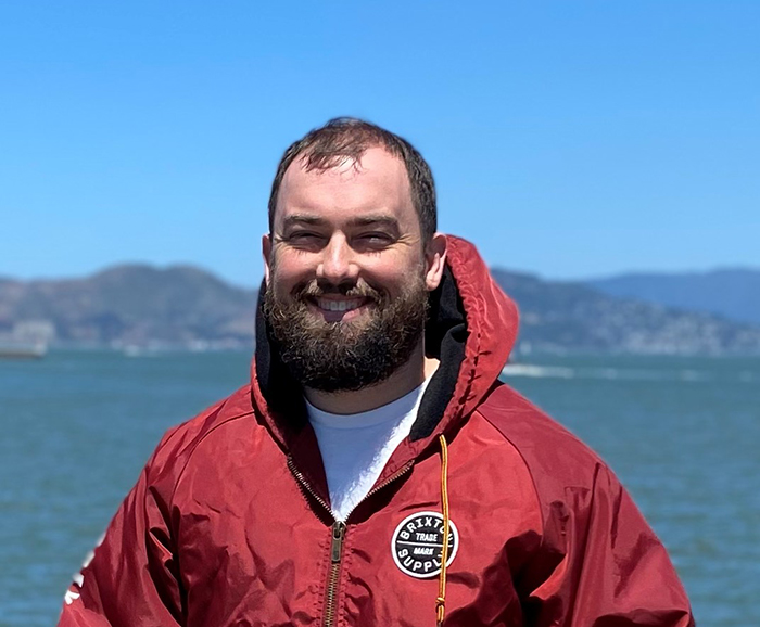 Portrait of a smiling, bearded young man wearing a red windbreaker, behind him lies a bay or lake.