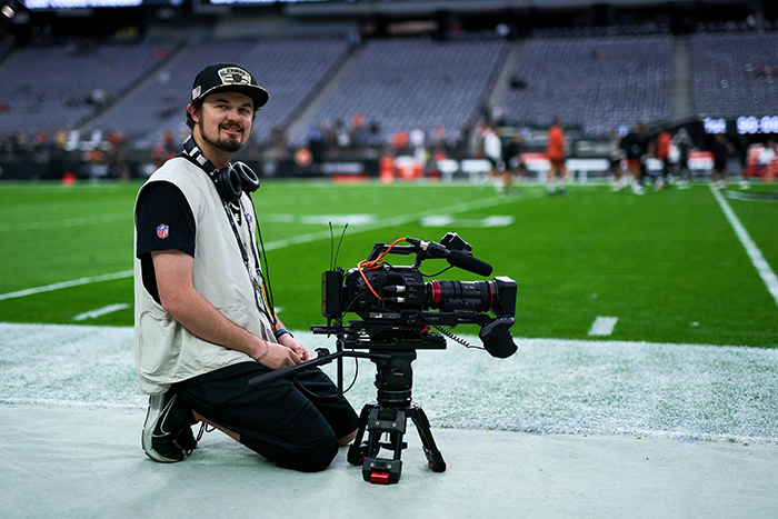 Bearded man in dark pants, tee shirt, and cap, light gray vest, kneels beside a video camera on a small tripod. He is on the sideline of a professional football field, with a practice in session and empty stands visible behind him.