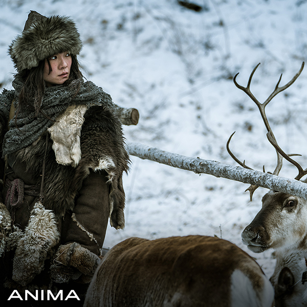 Central Asian woman, heavily dressed in furs, standing in a snowy landscape amongst reindeer